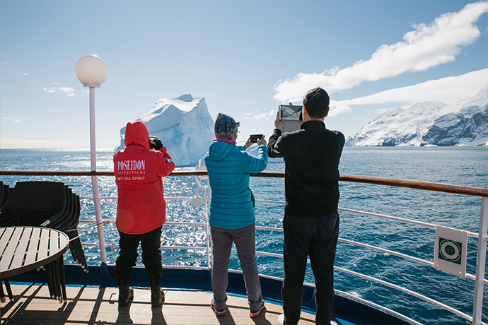 Gäste mit Handys uns Kameras an Deck des Expeditionskreuzfahrtschiffs fotografieren einen Eisberg