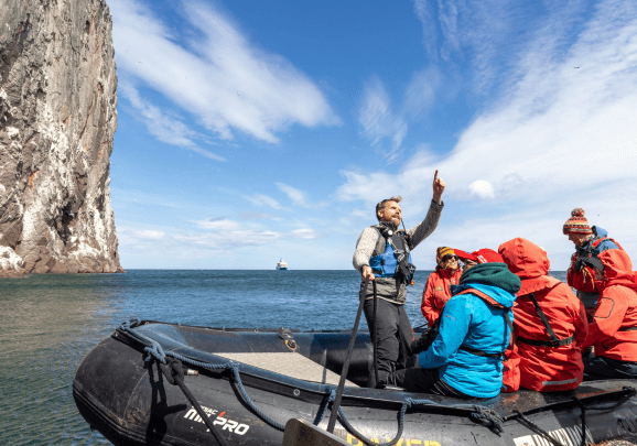 Im Schlauchboot steht der Expeditionsleiter und zeigt den Gästen die Basstölpel im Himmel vor der Insel Bass Rock