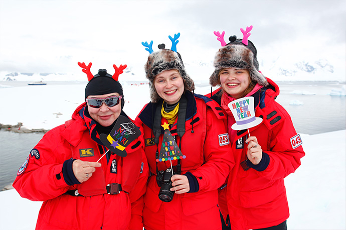 Drei personen in dicken Expeditions-Parkas feiern Silvester in der Antarktis. Dabei tragen Sie lustige Hüte und freuen sich.