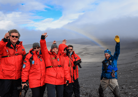 Auf der Insel Jan Mayen winken die Gäste der Kreuzfahrt und der Expeditionsleiter in die Kamera. Im Hintergrund ist ein Regenbogen zu sehen