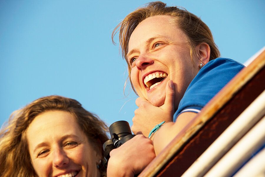 Zwei Frauen lachen am Oberdeck des Kreuzfahrtschiffs. Eine blickt in die Kamera und die zweite hält ein Fernglas in der Hand