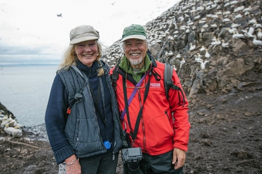 Zwei Gäste der Kreuzfahrt mit ihren Kameras auf der Insel Bass Rock mit vielen Basstölpeln der Vogelkolonie im Hintergrund