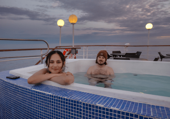 Zwei Kreuzfahrtgäste im Jacuzzi am Bord des Schiffs blicken in der Abenddämmerung in die Kamera.