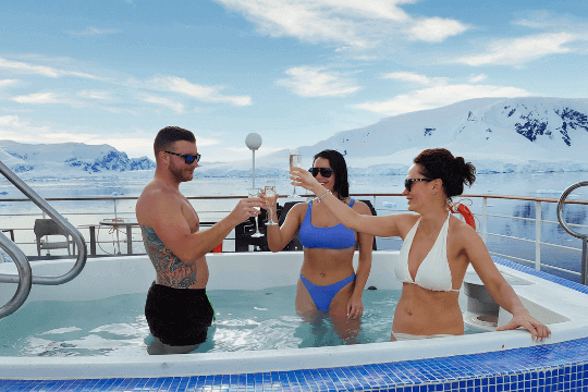 Drei Kreuzfahrtgäste stehen im Jacuzzi am Bord des Schiffs und prosten sich mit Getränken zu. Im Hintergrund ist die schneebedeckte Landschaft der Antarktis zu sehen.