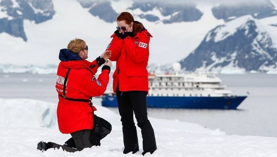 Ein Mann macht seiner Freundin in der Antarktis einen Heiratsantrag. Im Hintergrund ist das Kreuzfahrtschiff zu erkennen.