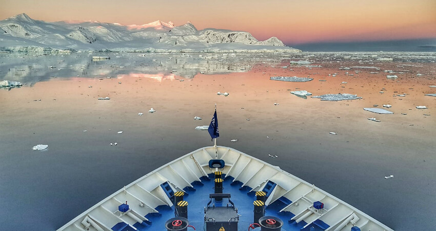 Der Bug vom Expeditionskreuzfahrtschiff steuert im Sonnenuntergang auf die schneebedeckte Küste der Antarktis zu. 7-Gruende01