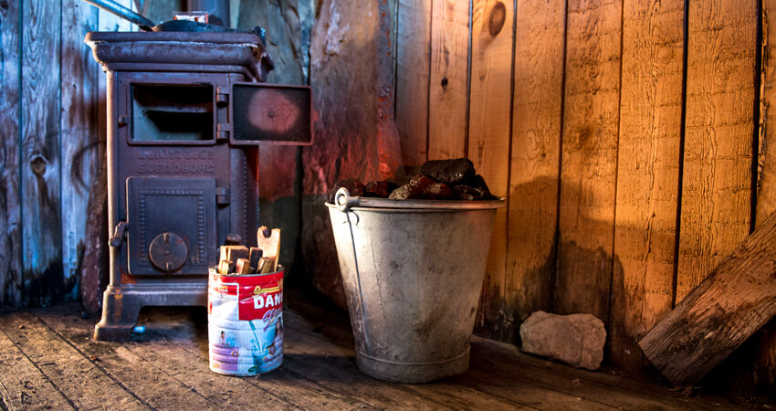 Ein alter Ofen in einer Holzhütte, davorstehend zwei Eimer mit Kohle und Zündstoff 07_Blomsterbukta_01_DSC8116_Anthony-Smith