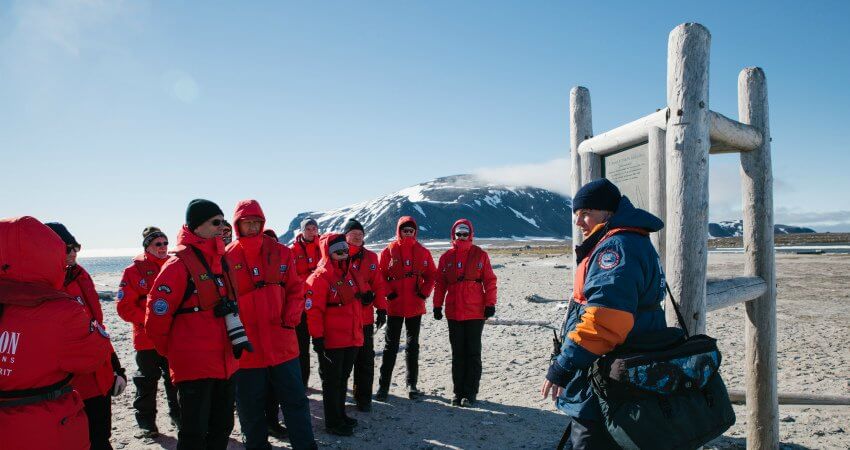 Auf Spitzbergen stehen Kreuzfahrtgäste mit ihrer Ausrüstung und Kameras bei dem Expeditionsleiter der ihnen eine Einweisung zu dem Gelände und der Geschichte gibt svalbard-4